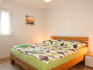 A bedroom with a wooden bed, a green and white patterned bedspread, a nightstand with a drawer, and a window with a view of a sunset.