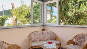 A corner room with two wicker chairs, a small round table with a white tablecloth, and a view of trees and buildings through large windows. - Apartment Mira by Interhome (Rab/Barbat)