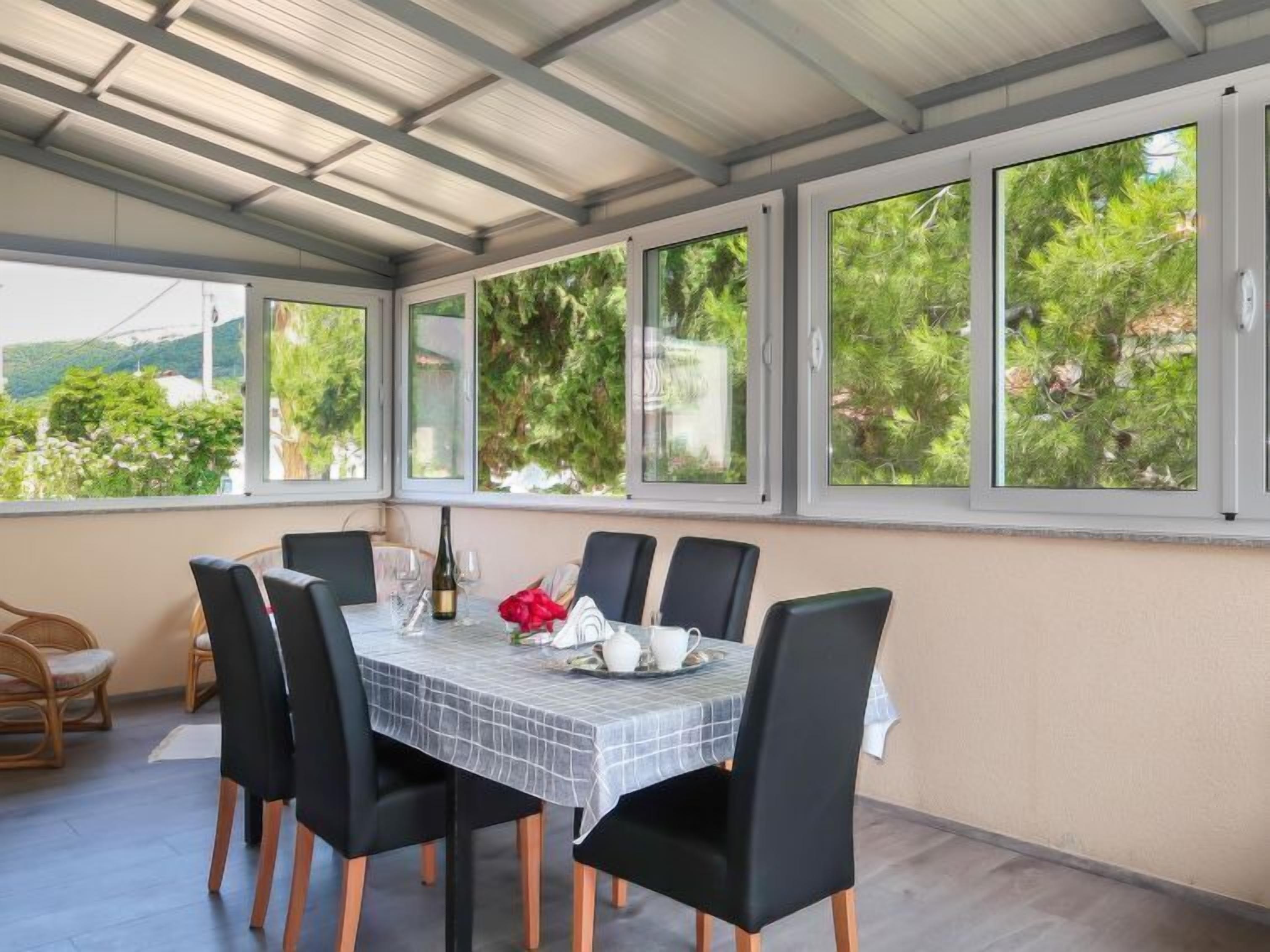 Ein überdachter Essbereich mit Tisch für vier Personen, umgeben von Stühlen, großen Fenstern mit Blick ins Grüne und einer Pergola.