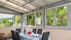 A covered dining area with a table set for four, surrounded by chairs, large windows with a view of greenery, and a pergola overhead. - Apartment Mira by Interhome (Rab/Barbat)