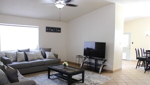 A living room with a grey sofa, a coffee table, a TV on a stand, and a dining area with chairs and a table. - Spacious Escape, 1KB, 2QB, Wifi - Private Yard (Albuquerque)