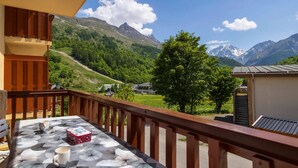 Ein Balkon mit Tisch, Tasse und einer Schachtel. Der Balkon bietet Blick auf eine gebirgige Landschaft mit Bäumen und Gebäuden.