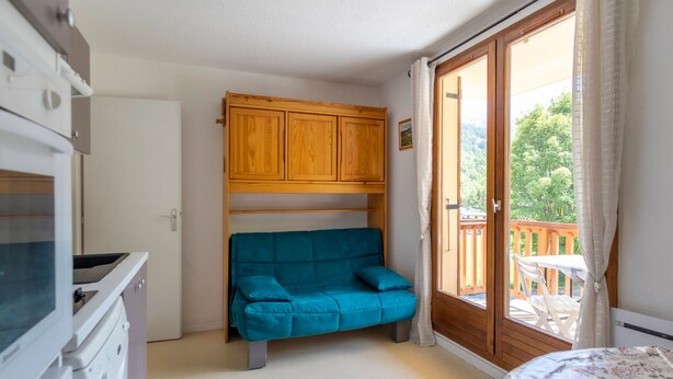 A room with a bed, a blue sofa, wooden cabinets, a washing machine, and a sliding glass door leading to an outdoor area with a table and chairs.