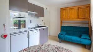 Eine kompakte Küche mit Geschirrspüler, Backofen und Mikrowelle. Ein blaugrünes Sofa steht an der Wand, und ein zartes Bettlaken mit Blumenmuster ist zu sehen.