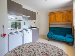 A compact kitchen with a dishwasher, oven, and microwave. A teal sofa is positioned against the wall, and a floral-patterned bedspread is visible.