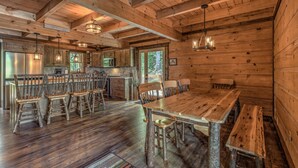 A rustic wooden cabin interior with a large dining table, wooden chairs, a kitchen area, and a bar.
