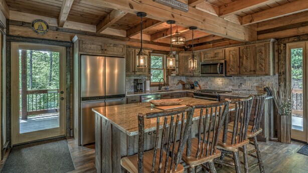 A rustic kitchen with wooden beams, a large island with bar stools, a refrigerator, a microwave, and a stone backsplash.