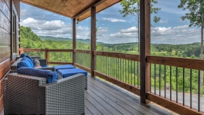 A covered outdoor seating area with wicker furniture and cushions, overlooking a lush green landscape.