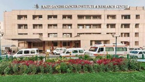 Het gebouw van het Rajiv Gandhi Cancer Institute & Research Centre met meerdere verdiepingen, een overdekte ingang en verschillende geparkeerde voertuigen ervoor.
