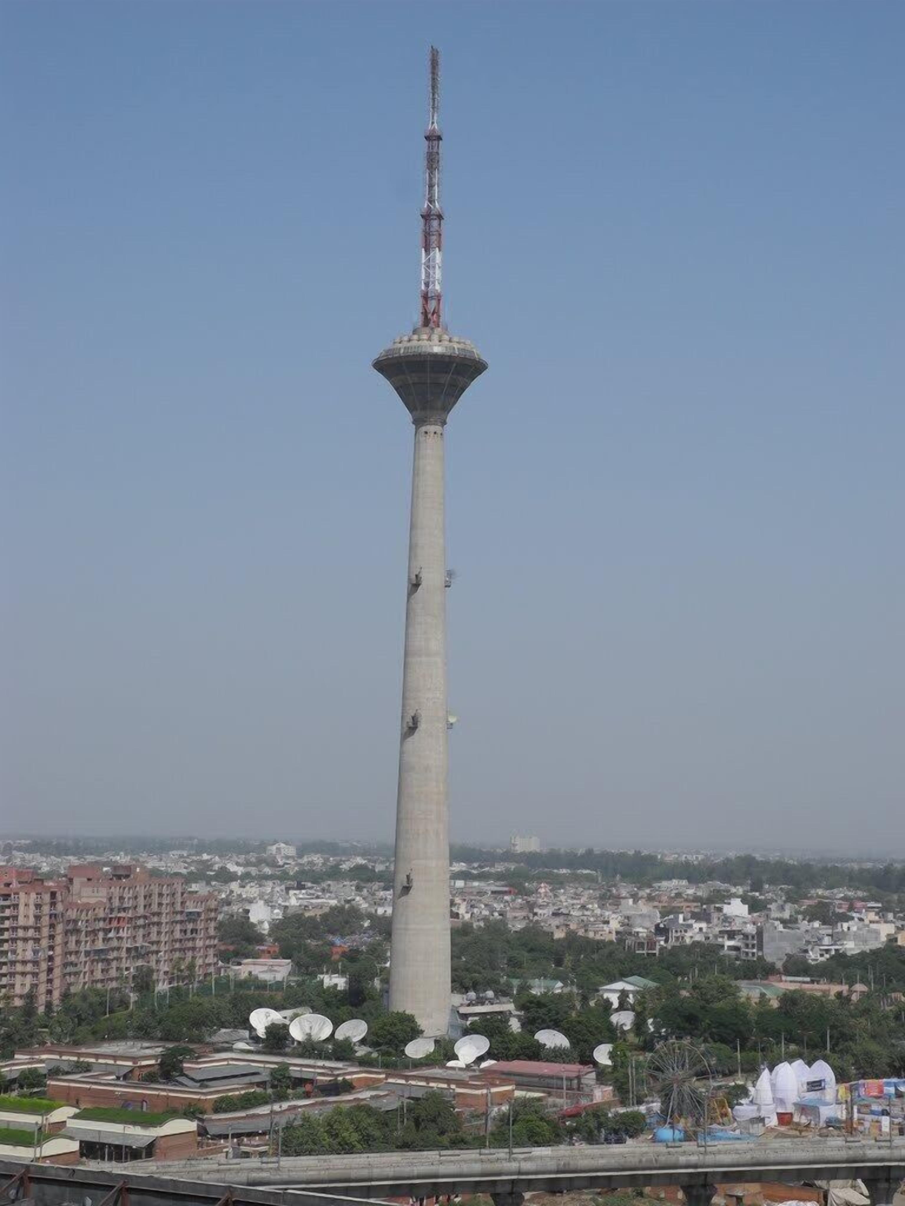 A tall tower with a cylindrical base and a pointed top, surrounded by a cityscape with buildings and greenery.