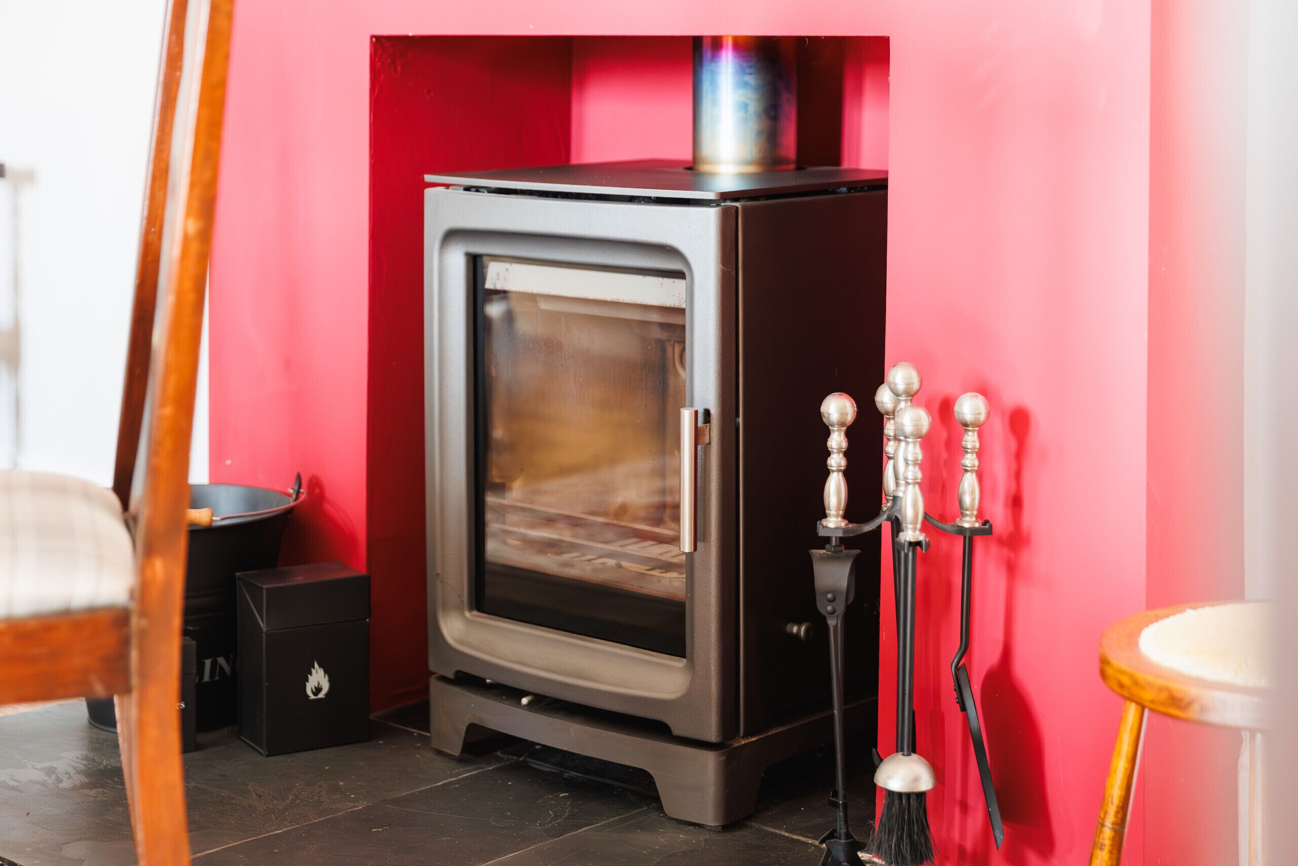 A compact wood-burning stove with a glass door, positioned against a red wall, accompanied by a black ashtray and a metal brush.