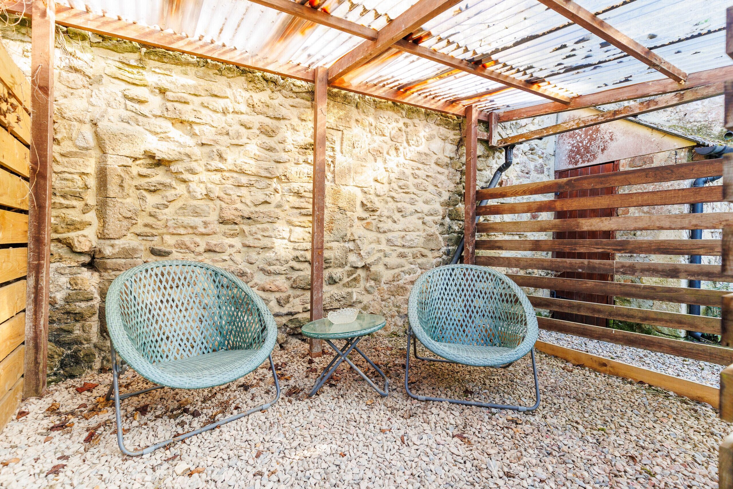 A covered outdoor seating area with two wicker chairs and a small table, set against a stone wall and wooden fence.