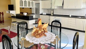 Un área de comedor con una mesa redonda de vidrio, mantel de encaje blanco y un centro de mesa con flores secas. Cuatro sillas negras rodean la mesa. Se observan armarios blancos y una encimera negra al fondo.