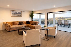 A modern living room with a sectional sofa, a round coffee table, and two armchairs. There is a large window with a view of a cityscape.