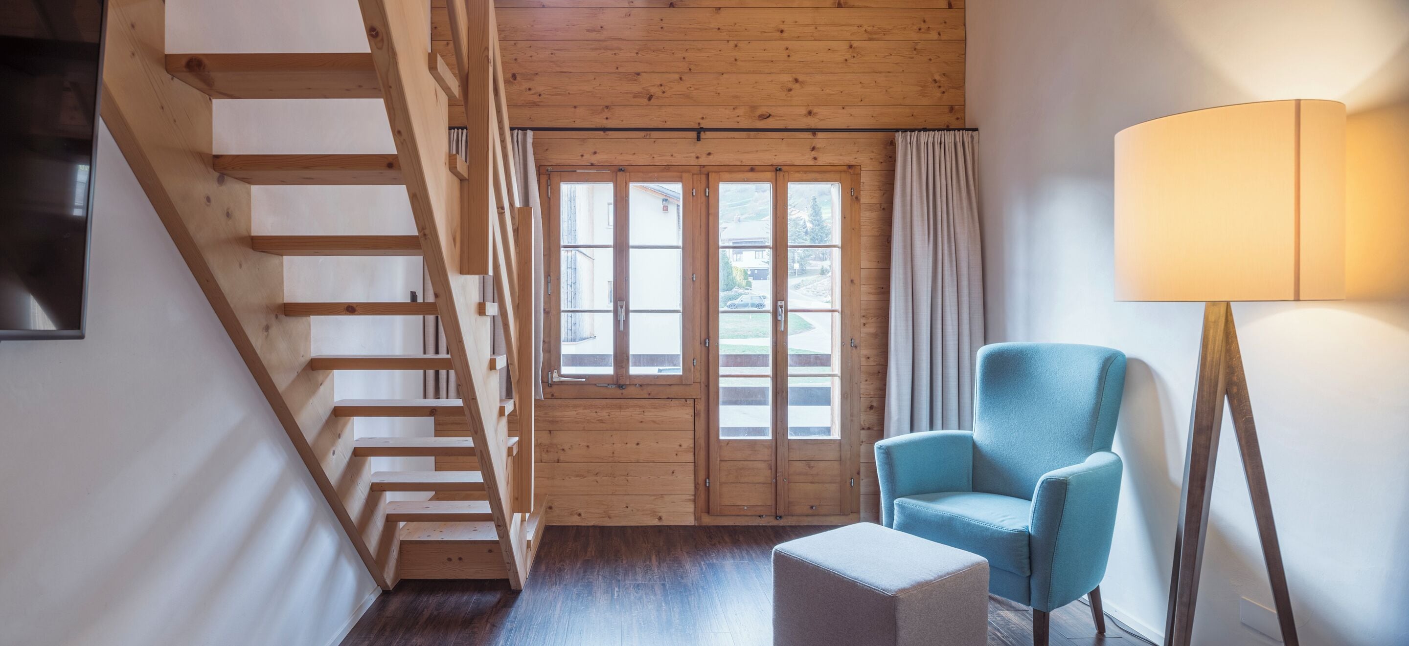 Ein Zimmer mit einer Holztreppe, einem blauen Sessel, einer Stehlampe und einem Fenster mit Vorhängen.