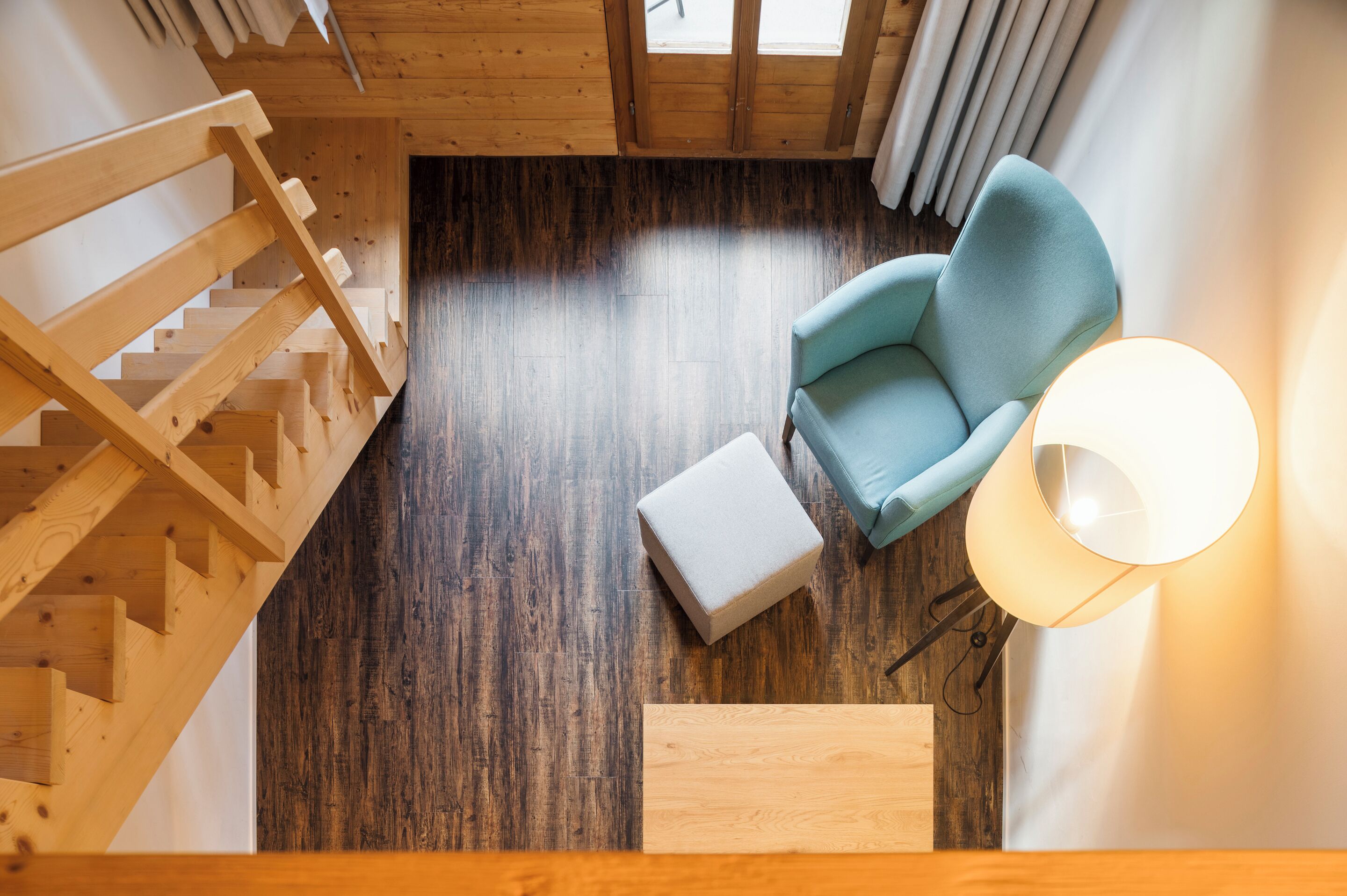 Una escalera de madera, un sillón azul claro, un otoman blanco y una lámpara de pie en una habitación con suelo de madera y techo inclinado.