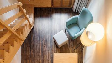 Una escalera de madera, un sillón azul claro, un otoman blanco y una lámpara de pie en una habitación con suelo de madera y techo inclinado.