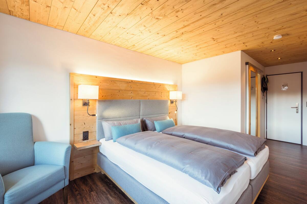 Ein modernes Schlafzimmer mit Holzboden, einem großen Bett mit grauer Bettwäsche, einem blauen Sessel und Nachttischlampen.