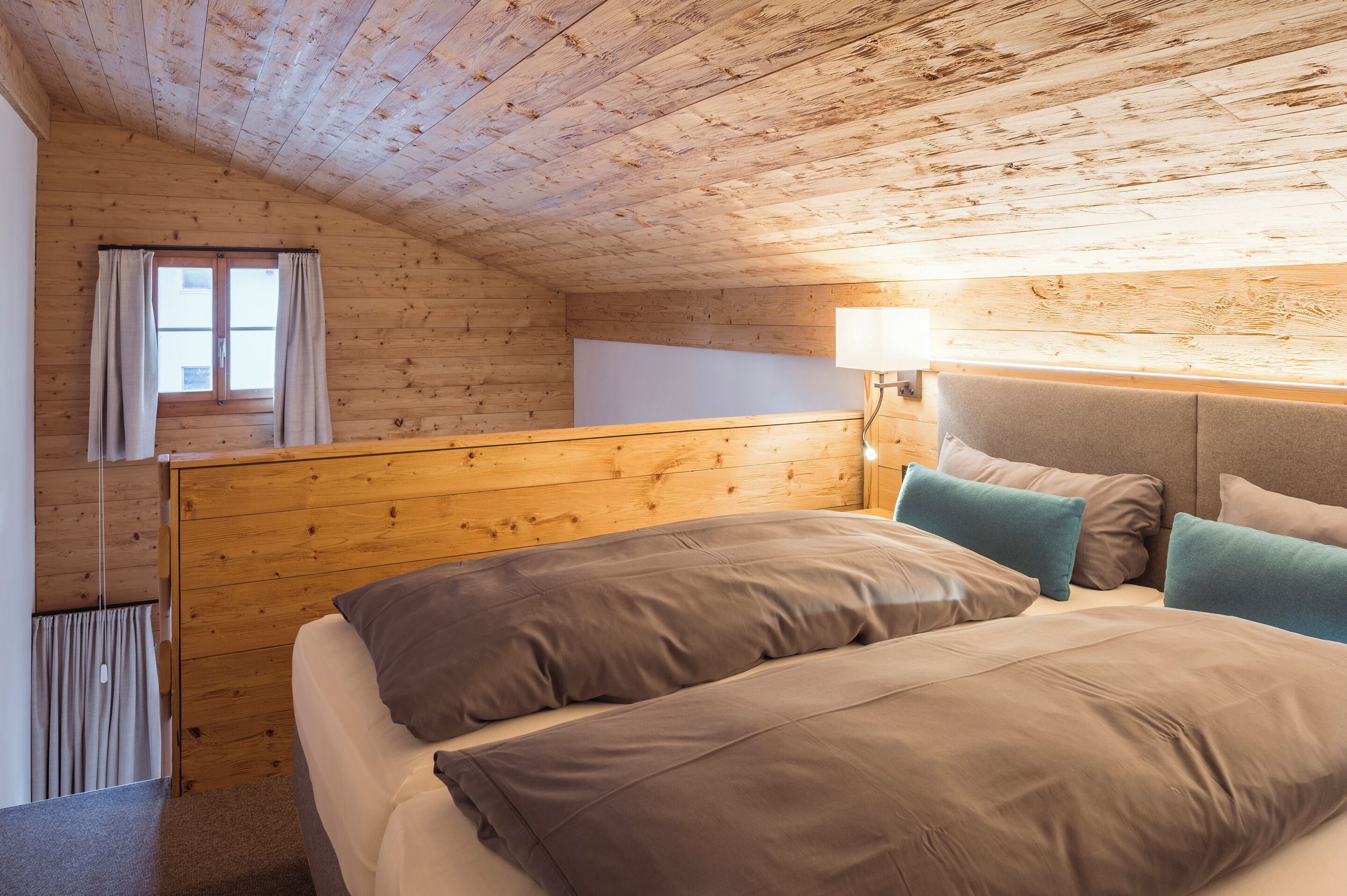 Een kamer met houten lambrisering, een bed, een raam met gordijnen en een wandlamp.