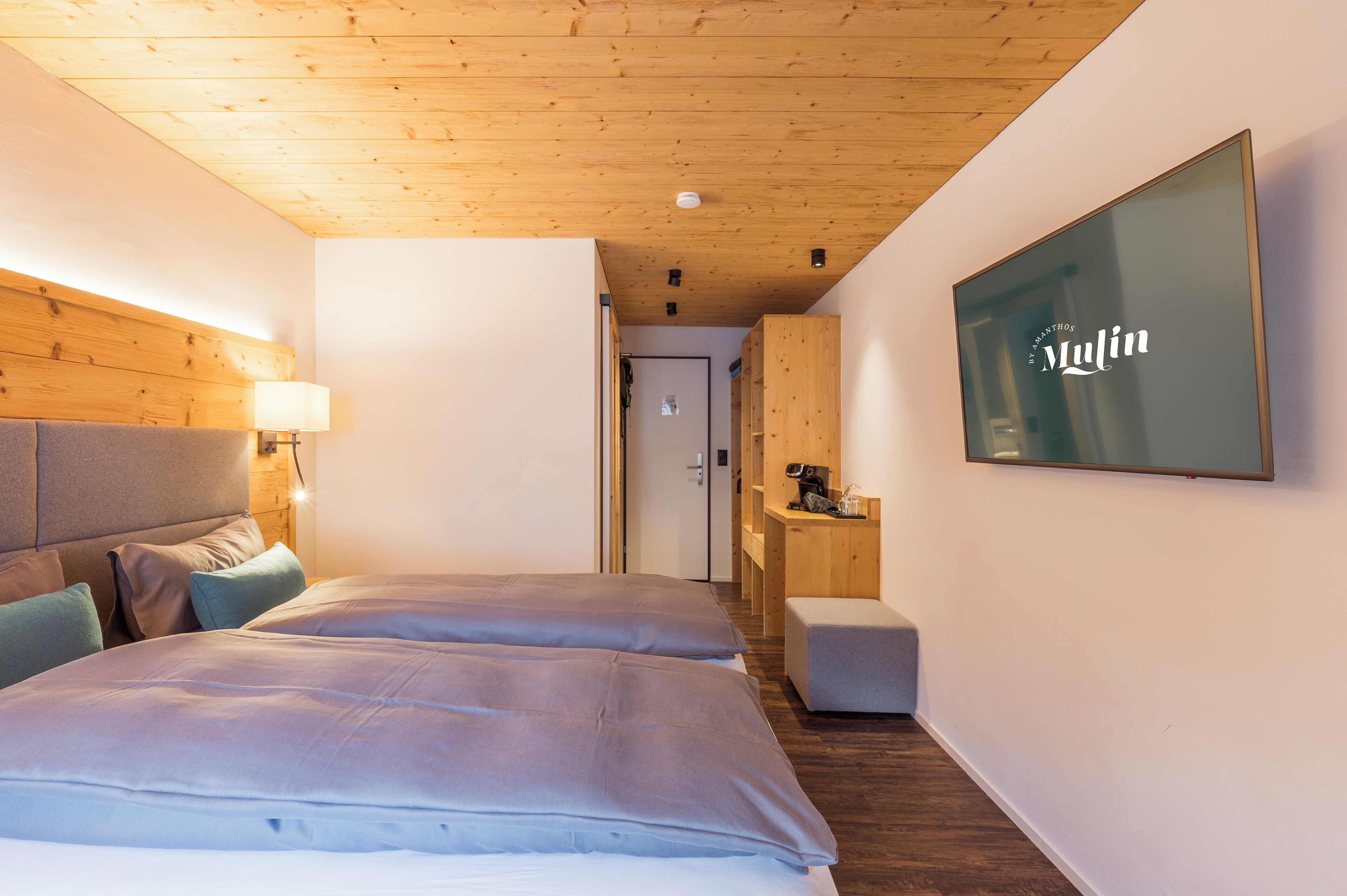 Ett modernt hotellrum med ett träpanelat tak, en stor säng med grå sängkläder, en liten sittgrupp och en väggmonterad tv som visar hotellets namn.