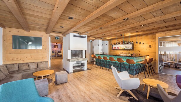 A modern lounge area with wooden ceilings, a bar with stools, and a fireplace. - Hotel Mulin by Amanthos (Brigels)