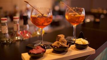 Two glasses of amber-colored cocktails with a wooden tray containing a small bowl of food and a bottle of sauce.