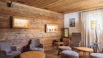 A cozy room with wooden walls, a large chair, ottomans, a small table, and a lamp.
