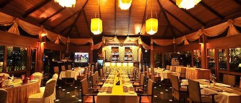 A restaurant with a long table layout, chandeliers, and a bar area.