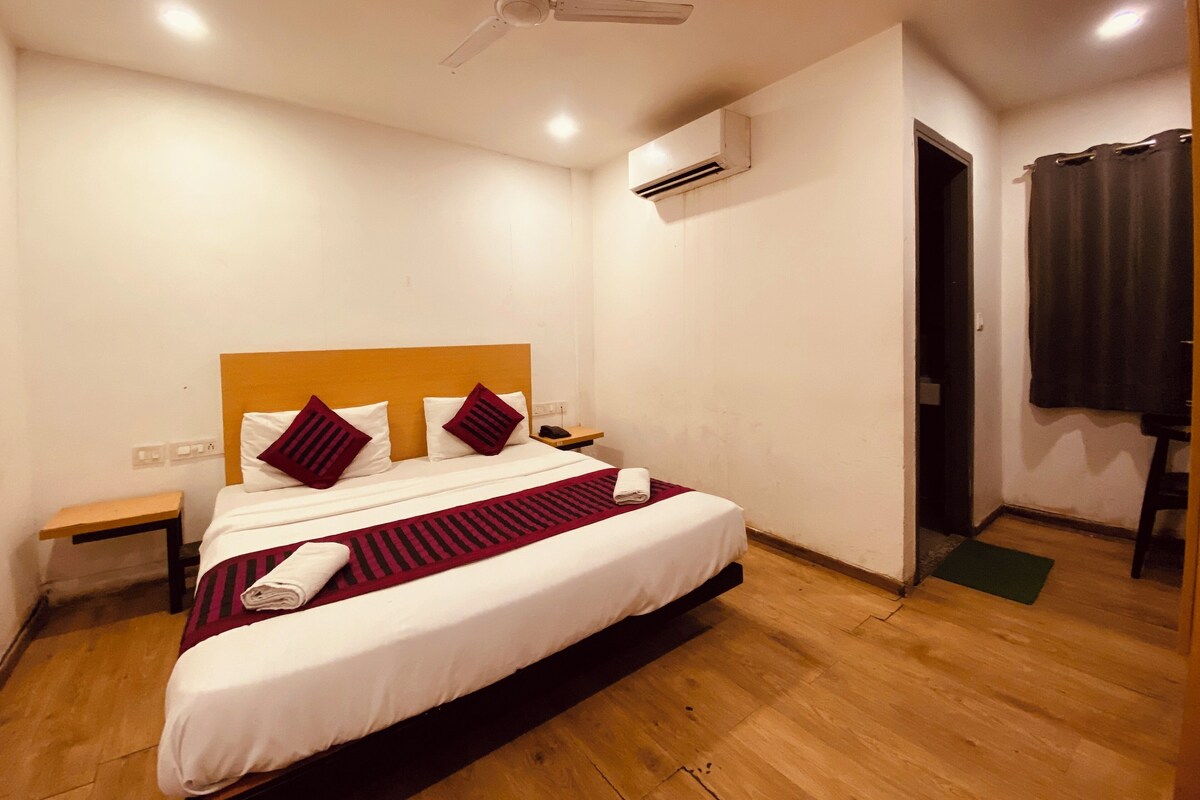 A neatly made bed with white and maroon bedding, a wooden headboard, and a red striped runner. There is a ceiling fan, an air conditioning unit, and a small bedside table with a lamp.