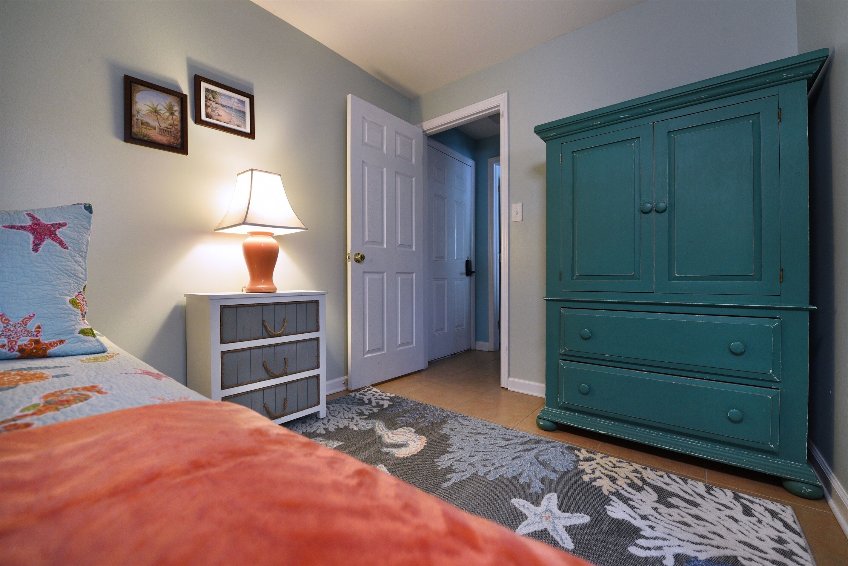 Un dormitori amb un llit, una tauleta de nit amb una làmpada, un armari i una porta que condueix a una altra habitació.