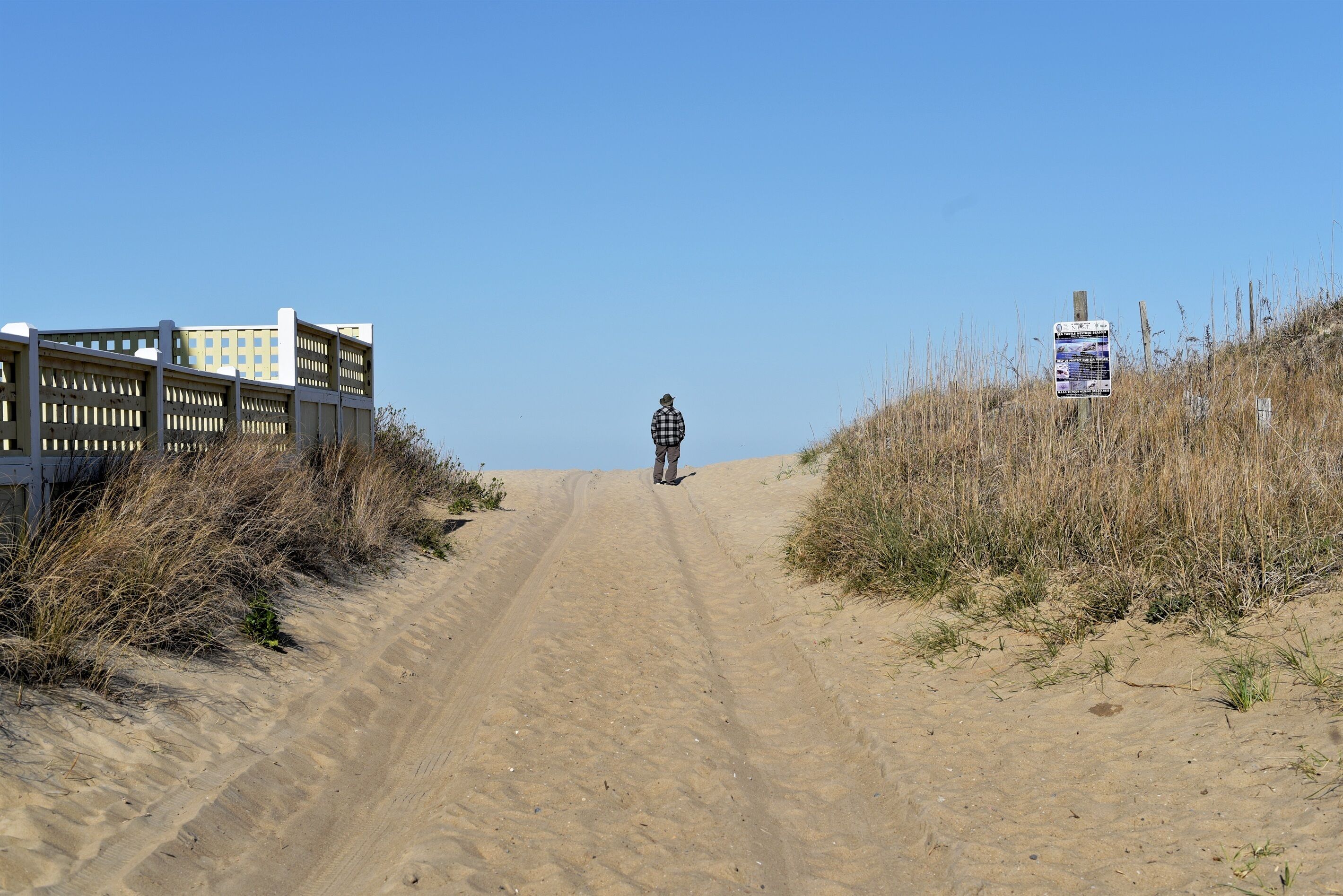 Un chemin de sable mène à un bâtiment, avec une clôture d’un côté et un panneau de signalisation de l’autre.