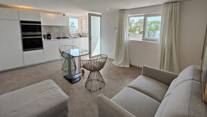 A modern kitchen with built-in appliances, a dining area with a glass table and metal chairs, and a large window with curtains.