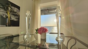 A room with a glass table, two crystal candle holders with candles, a vase with pink flowers, and a painting on the wall.