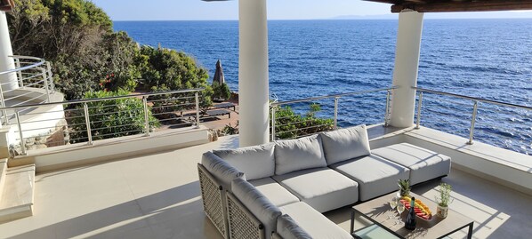 Un balcon avec un canapé blanc, une table en verre sur laquelle sont disposés des fruits et une bouteille, et une vue sur la mer.