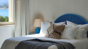 Un lit avec une tête de lit bleue, une literie blanche et plusieurs oreillers. Une lampe se trouve sur une table de chevet à côté du lit. Une fenêtre avec des rideaux est visible, offrant une vue sur la mer et les collines.