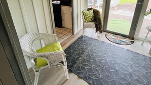 Una habitación con una silla de mimbre blanca, un tapete gris estampado y vistas a una cocina y zona exterior a través de una puerta corredera.