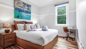 Ein Schlafzimmer mit einem hölzernen Bett, einem Nachttisch, einem Schreibtisch und einem Fenster mit Blick auf Bäume.