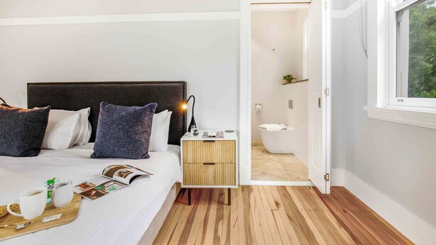 Ein modernes Schlafzimmer mit Holzboden, einem Bett mit weißer Bettwäsche und dunklem Kopfteil, einem Nachttisch mit Lampe sowie Blick ins Badezimmer durch eine geöffnete Tür.