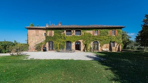 Un grande edificio in pietra con tetto a tegole, muri ricoperti d'edera e vialetto sterrato che conduce a una panchina di legno.