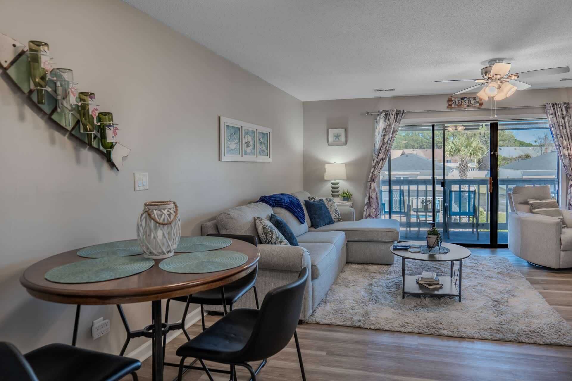 A living room with a sofa, coffee table, and chairs, a dining area with a table and chairs, and a view through a glass door to the outside.