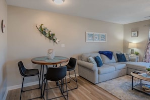 Una sala de estar con un sofá, dos sillones, una mesa redonda con un jarrón decorativo, obras de arte en la pared y una lámpara.