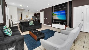 Un salon moderne avec un canapé noir, un fauteuil gris et une table basse. Un téléviseur à écran plat est fixé au mur, et on aperçoit un coin cuisine au fond de la pièce.