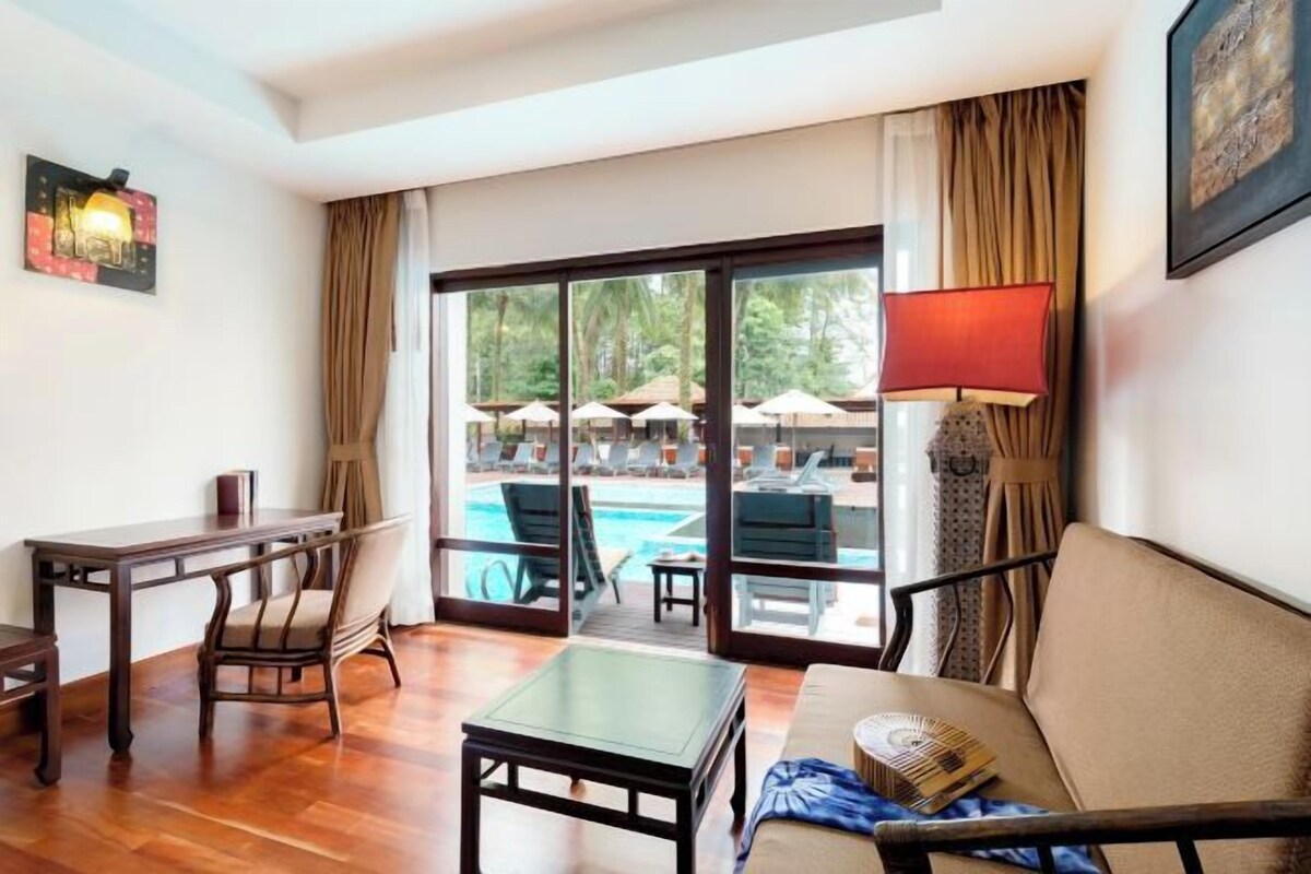 Ein Hotelzimmer mit Holzboden, einer Couch, einem kleinen Tisch, einem Sessel, einem Schreibtisch und Blick auf einen Pool mit Sonnenschirmen.