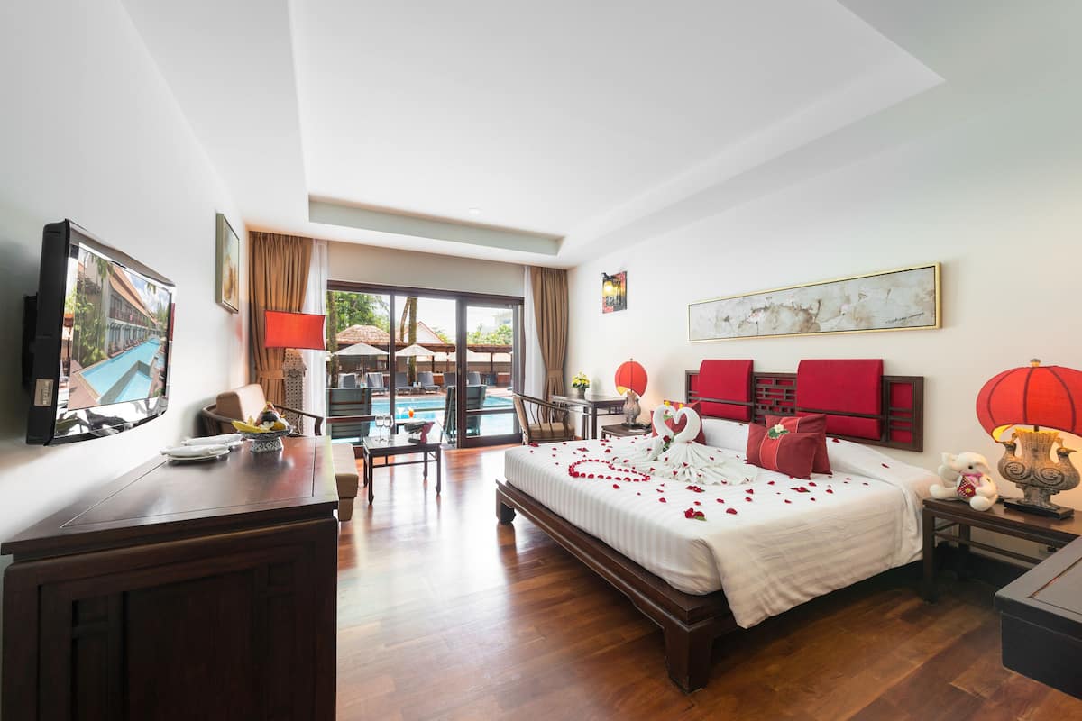 Quarto de hotel com cama, escrivaninha, cadeira, televisão, luminária, abajur vermelho, vaso, mesa pequena, travesseiro vermelho, travesseiro branco, decoração em formato de coração vermelho, lanterna vermelha, pintura e janela com cortinas.