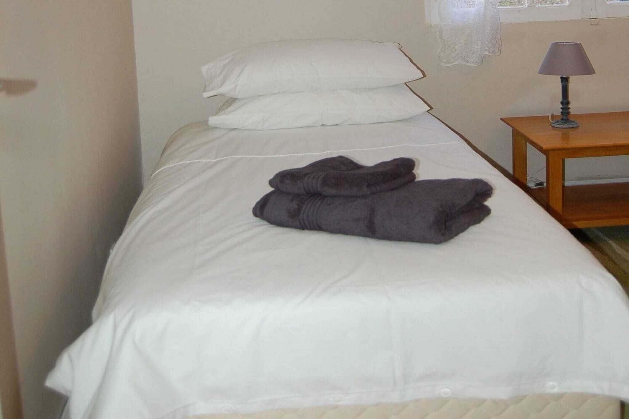 A single bed with white bedding, a folded gray towel, and a wooden nightstand with a lamp.