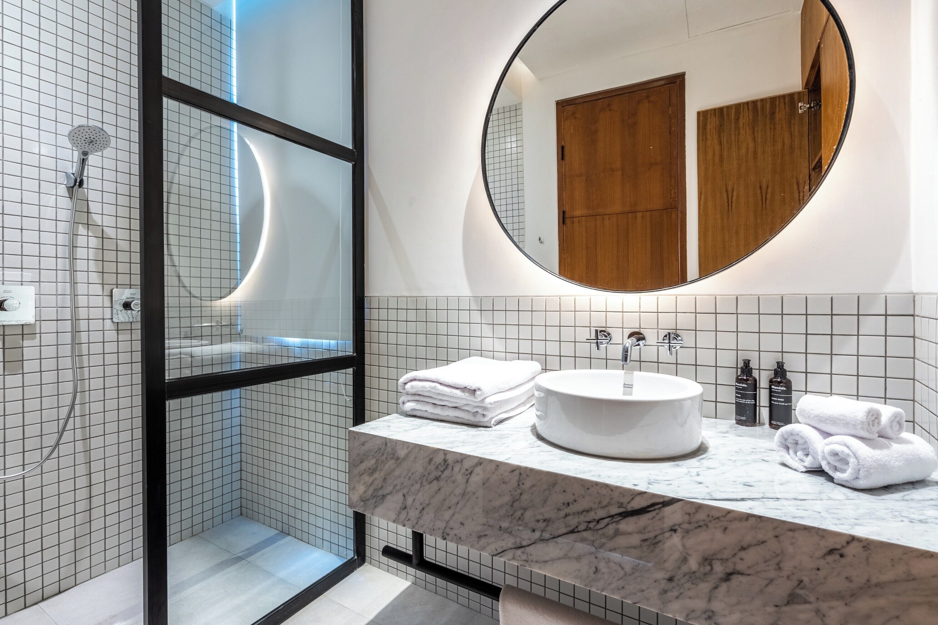 A modern bathroom with a glass-enclosed shower, a round mirror, and a marble countertop with a sink and towels.