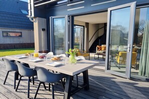 Un espace repas extérieur moderne, avec une table dressée pour deux personnes, entourée de chaises, et une porte vitrée donnant sur un espace de détente abrité.