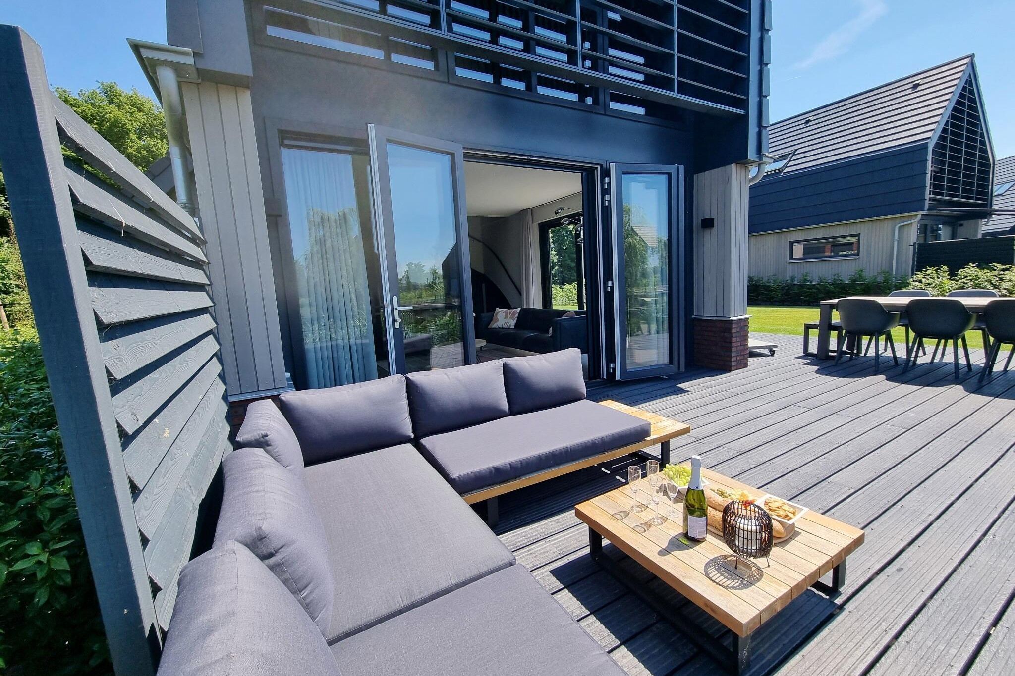 A modern outdoor deck with a sofa and a table set for dining, featuring a bottle of wine and a bowl of fruit.