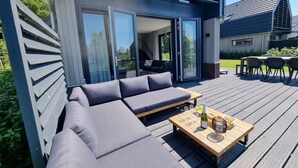 A modern outdoor deck with a sofa and a table set for dining, featuring a bottle of wine and a bowl of fruit. - Water Villa in Balk with Lake Access (Balk)