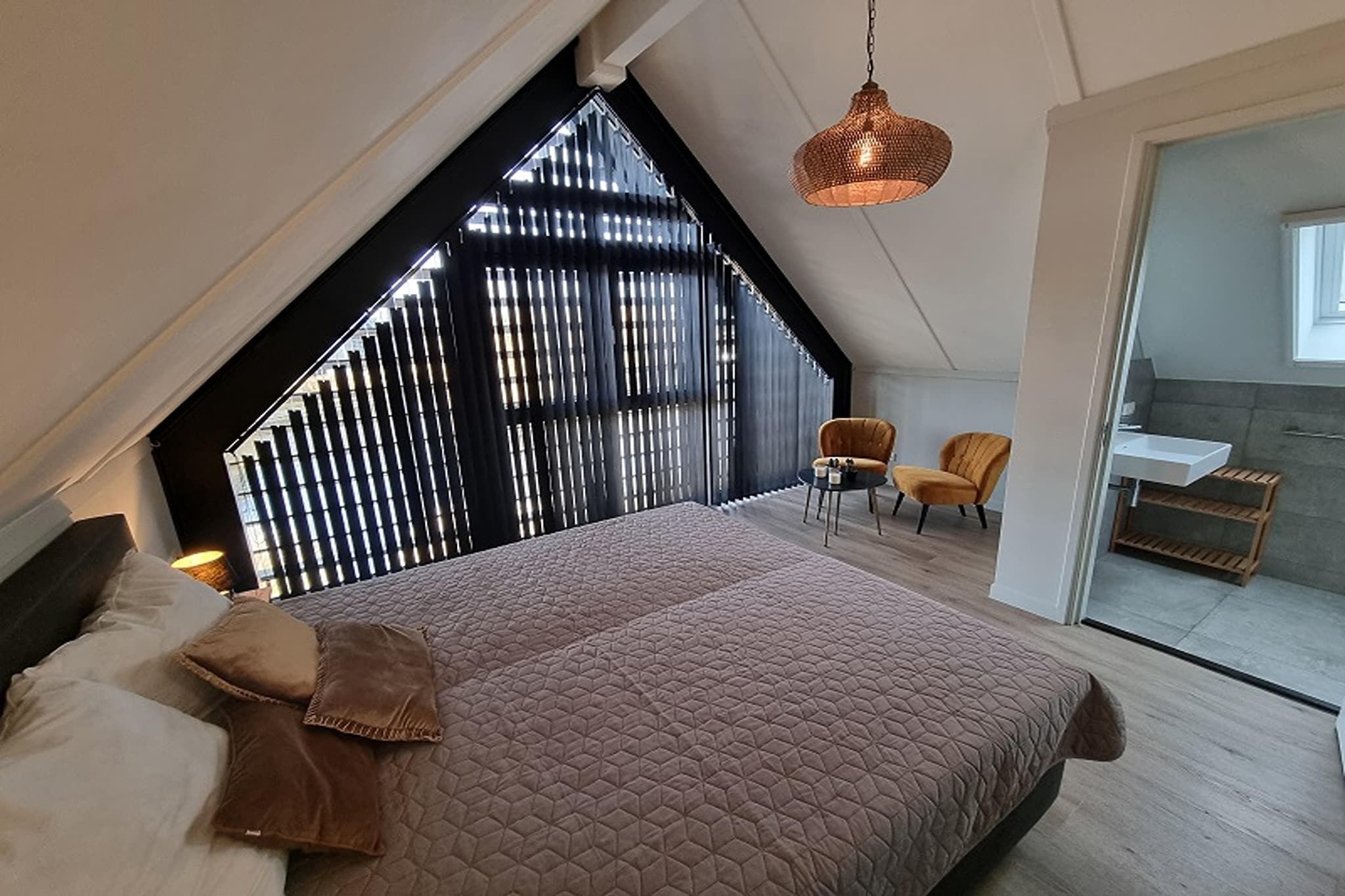 A bedroom with a large bed, a chair, a hanging light fixture, and a view of a kitchen.